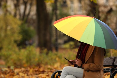 Photo of Woman with rainbow umbrella and smartphone sitting on bench in autumn park, space for text