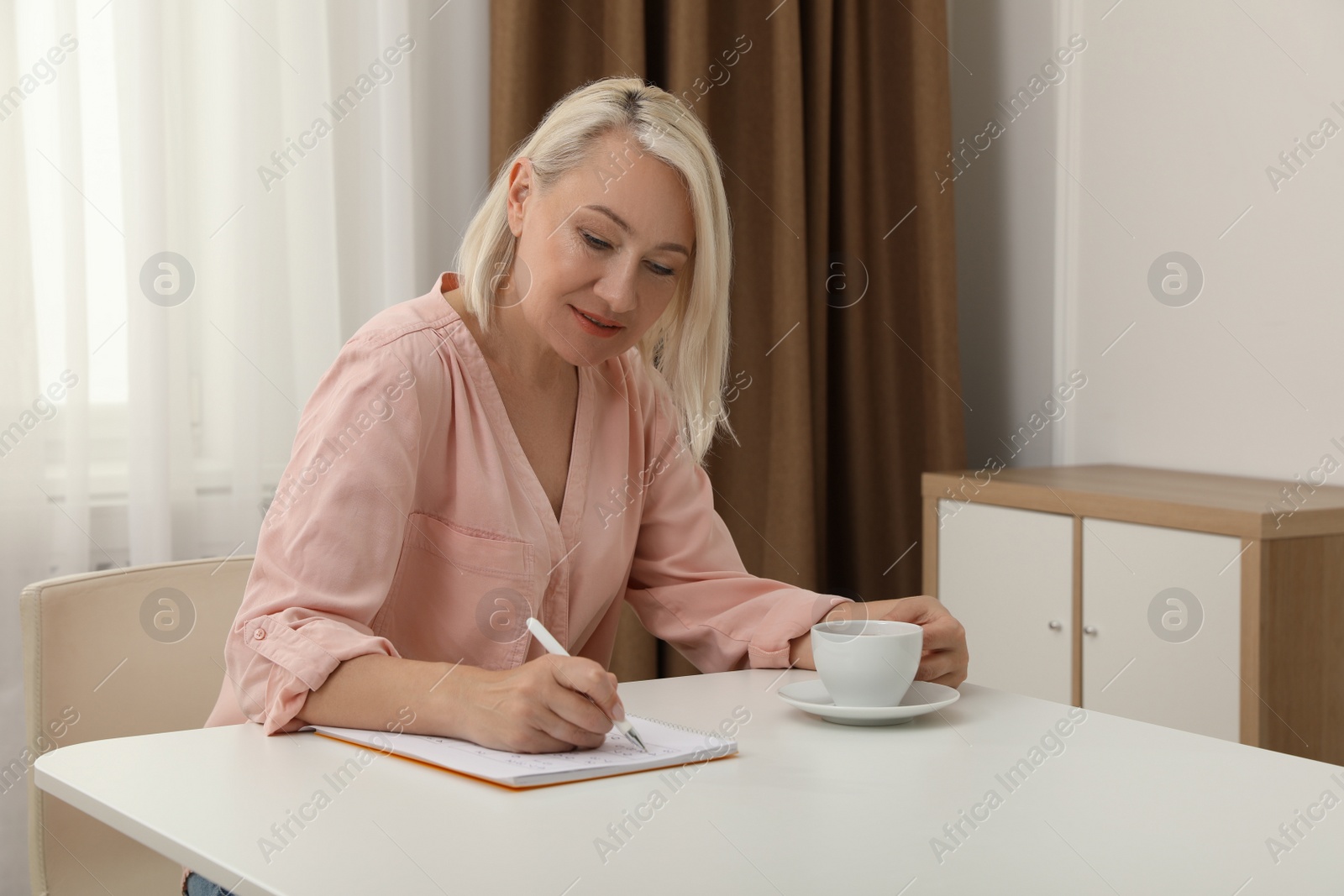 Photo of Middle aged woman with cup of drink solving sudoku puzzle at table indoors