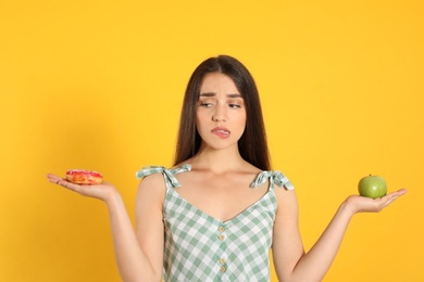 Doubtful woman choosing between apple and doughnut on yellow background