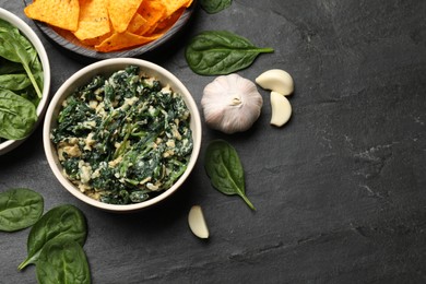 Photo of Tasty spinach dip with egg in bowl, garlic and nachos chips on black table, flat lay. Space for text