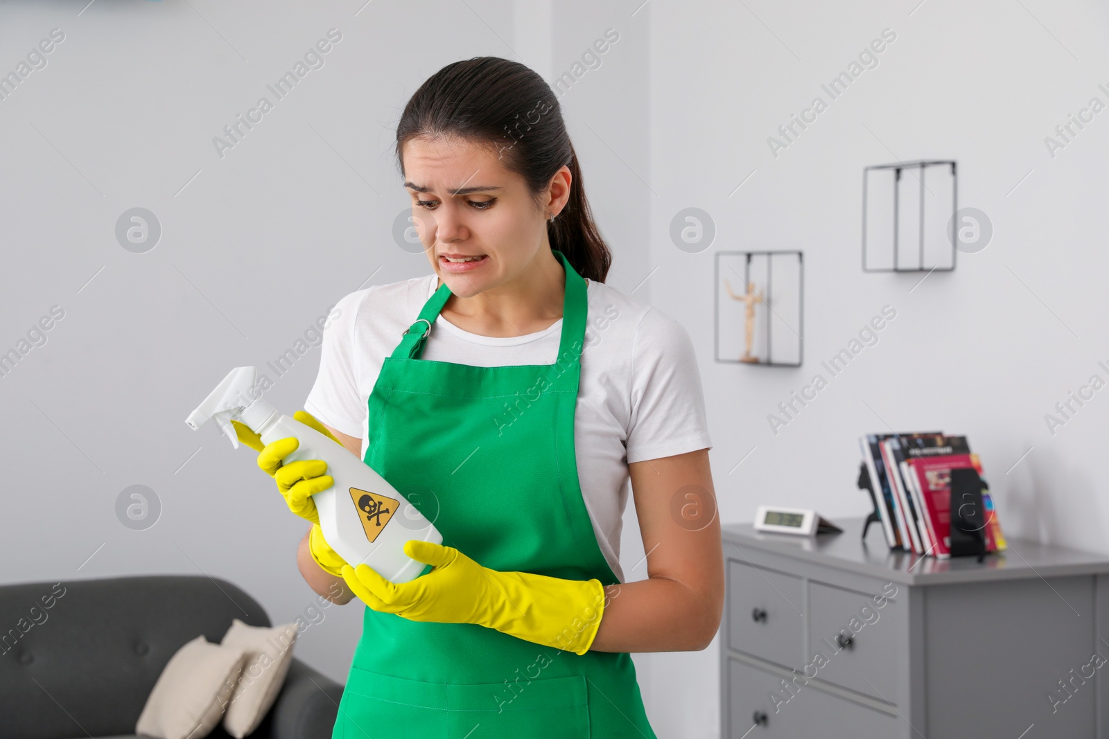 Photo of Woman looking at bottle of toxic household chemical with warning sign indoors