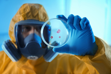 Photo of Scientist in chemical protective suit with Petri dish at laboratory, focus on hand. Virus research