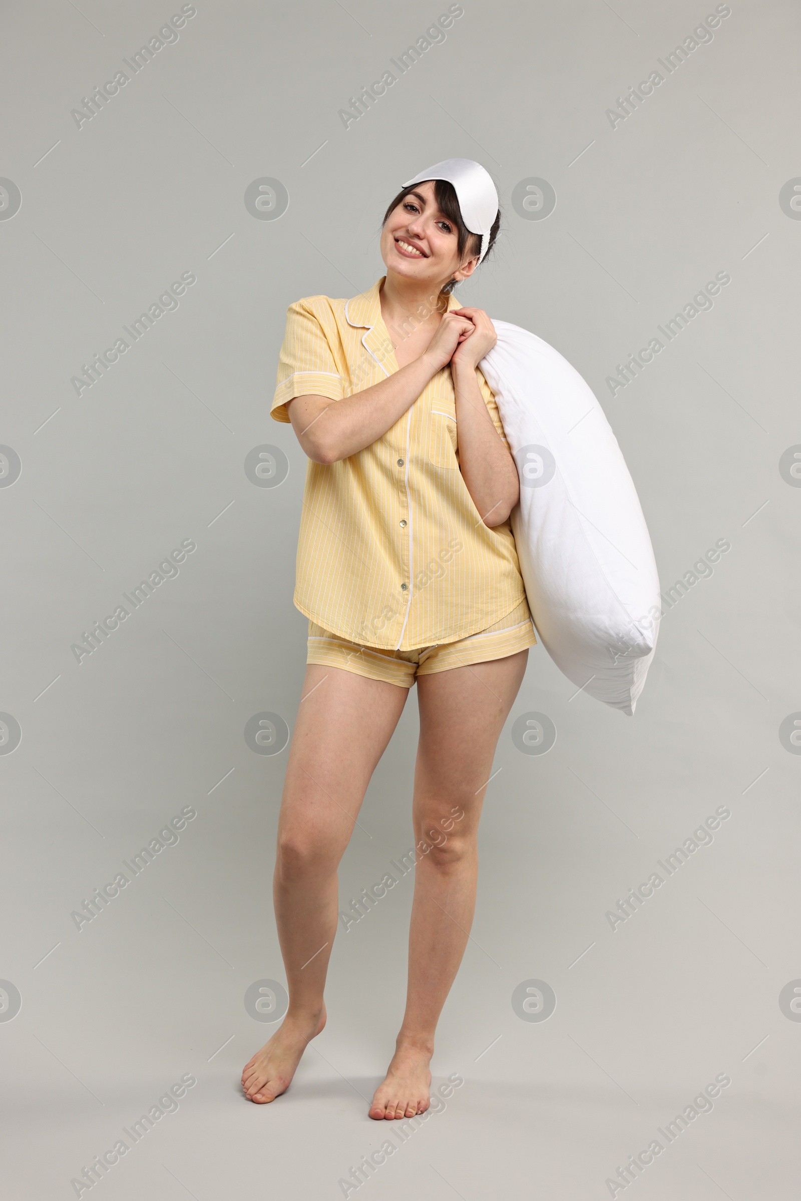 Photo of Happy woman in pyjama and sleep mask holding pillow on grey background