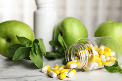 Photo of Bottle with vitamin pills, apples and mint on white marble table