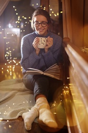 Woman with cup of hot beverage reading book at home in winter evening