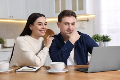 Photo of Happy couple with laptop shopping online at wooden table in kitchen