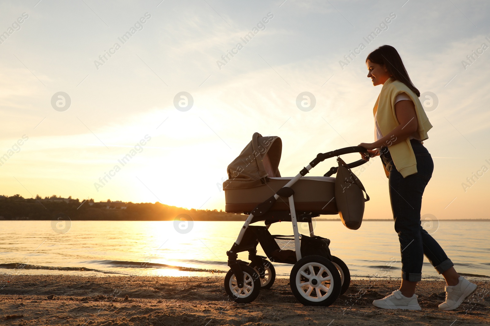 Photo of Happy mother with baby in stroller walking near river at sunset