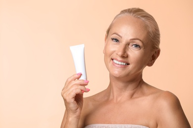 Portrait of beautiful mature woman with perfect skin holding tube of cream on beige background. Space for text