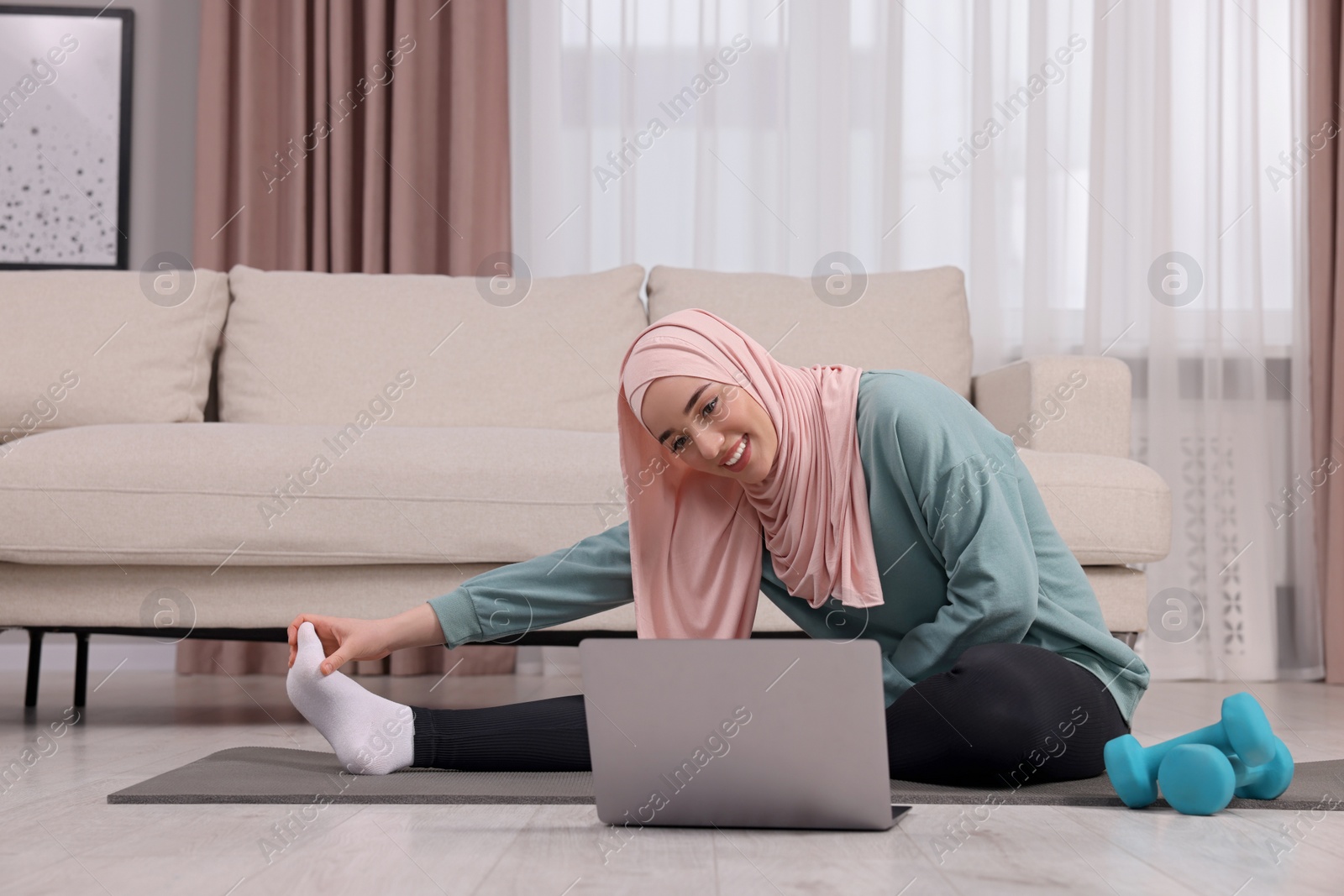 Photo of Muslim woman in hijab stretching near laptop on fitness mat at home