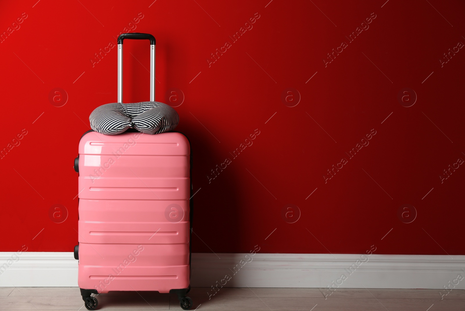 Photo of Pink suitcase and striped travel pillow near red wall, space for text