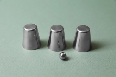 Photo of Three metal cups and ball on pale olive background. Thimblerig game