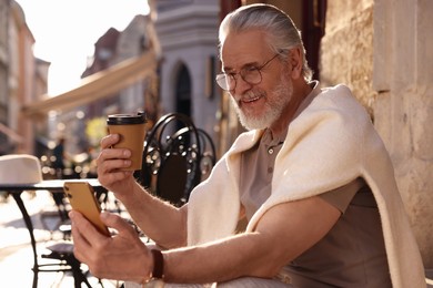 Handsome senior man sitting on doorstep, using smartphone and drinking coffee outdoors, space for text