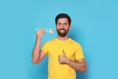 Photo of Happy man holding sushi roll with chopsticks and showing thumbs up on light blue background