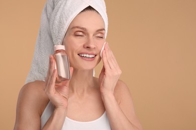 Photo of Beautiful woman in terry towel removing makeup with cotton pad on beige background, space for text