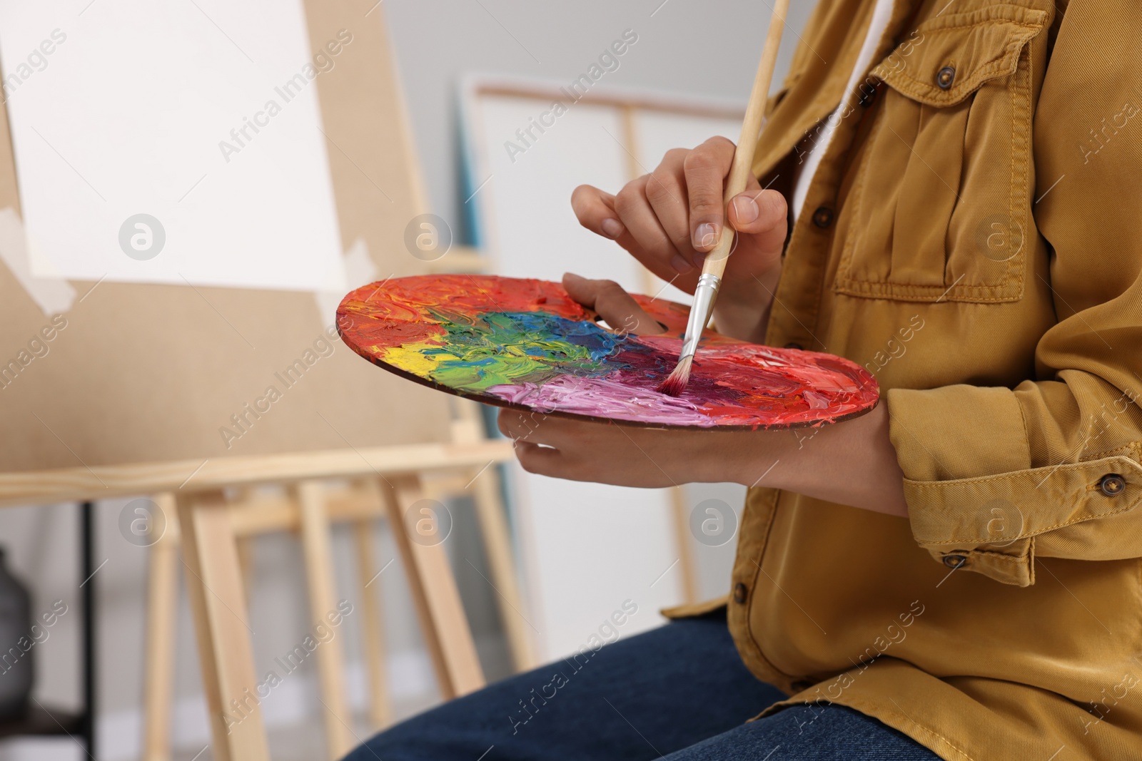 Photo of Woman mixing paints on palette with brush near easel in studio, closeup