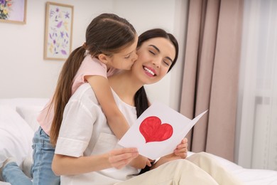 Photo of Happy woman with her daughter and handmade greeting card on bed at home. Mother's day celebration