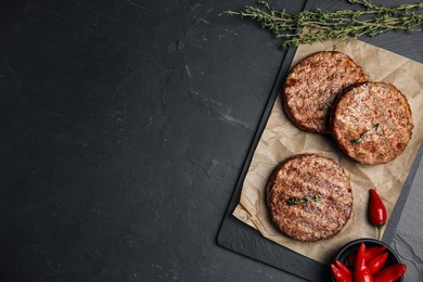 Tasty grilled hamburger patties, thyme and chili peppers on black table, flat lay. Space for text