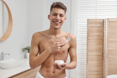 Photo of Handsome man applying moisturizing cream onto his shoulder in bathroom