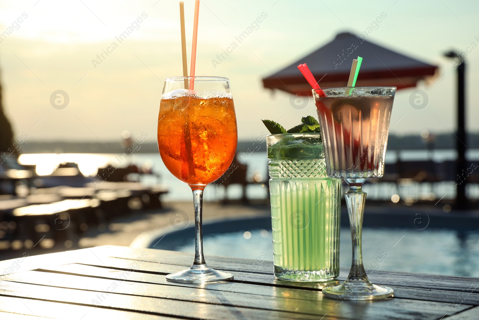 Photo of Glasses of fresh summer cocktails on wooden table near swimming pool outdoors at sunset