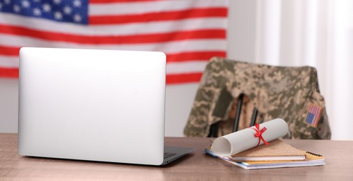 Image of Military education. Laptop, notebooks and diploma on wooden table, chair with soldier's jacket against flag of United States indoors