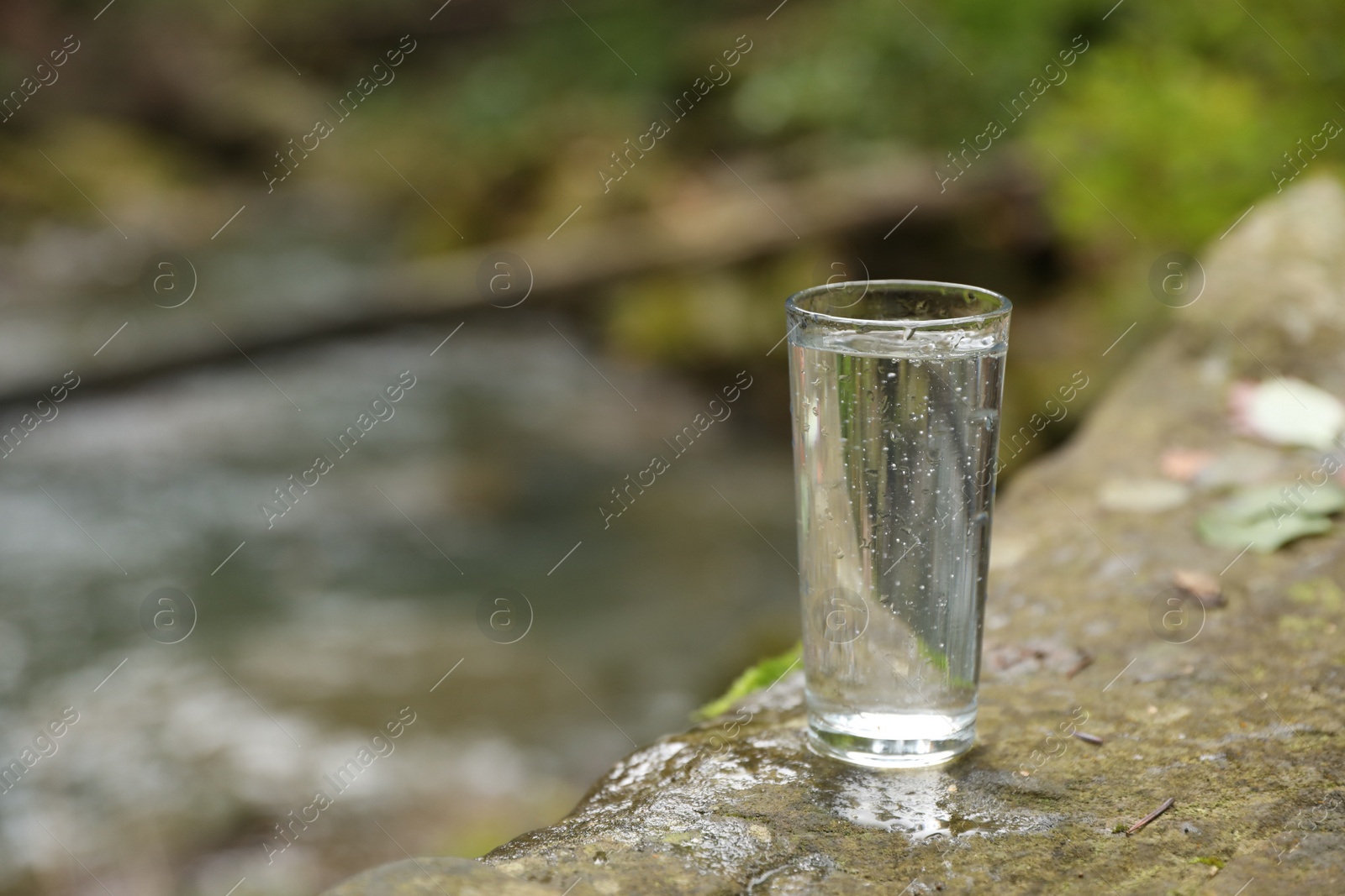 Photo of Glass of fresh water on stone near stream. Space for text