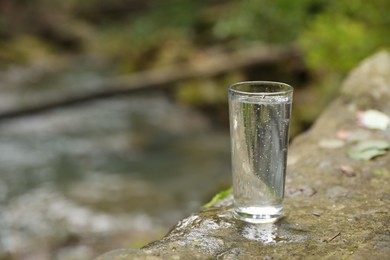 Photo of Glass of fresh water on stone near stream. Space for text