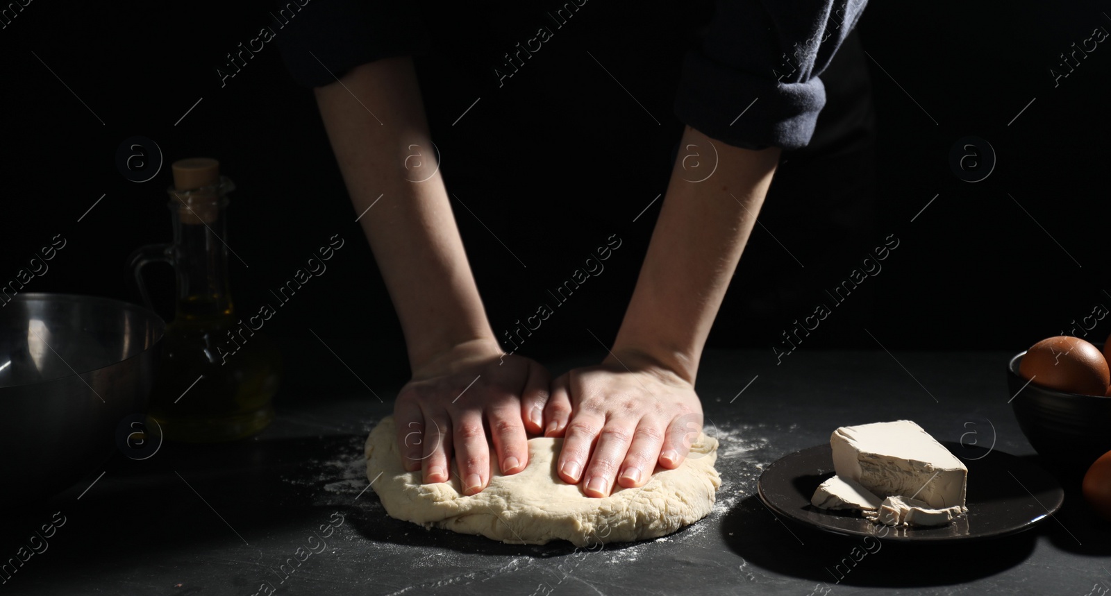 Photo of Making bread. Woman kneading dough at black table on dark background, closeup