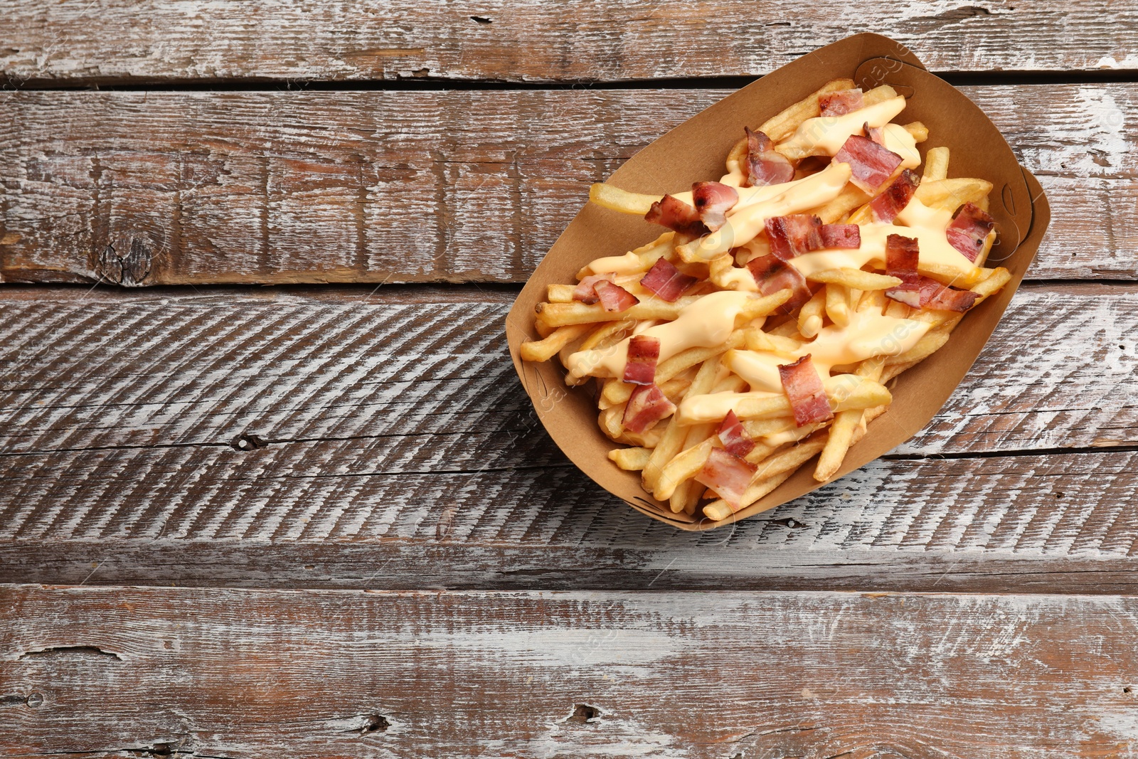 Photo of Tasty potato fries, cheese sauce and bacon in paper container on wooden table, top view. Space for text