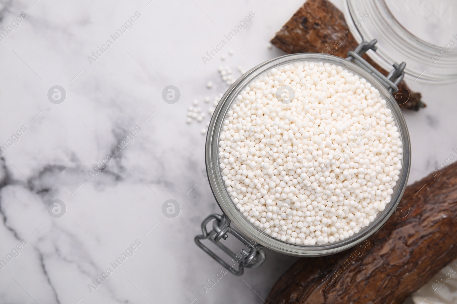 Photo of Tapioca pearls in jar and cassava roots on white marble table, flat lay. Space for text