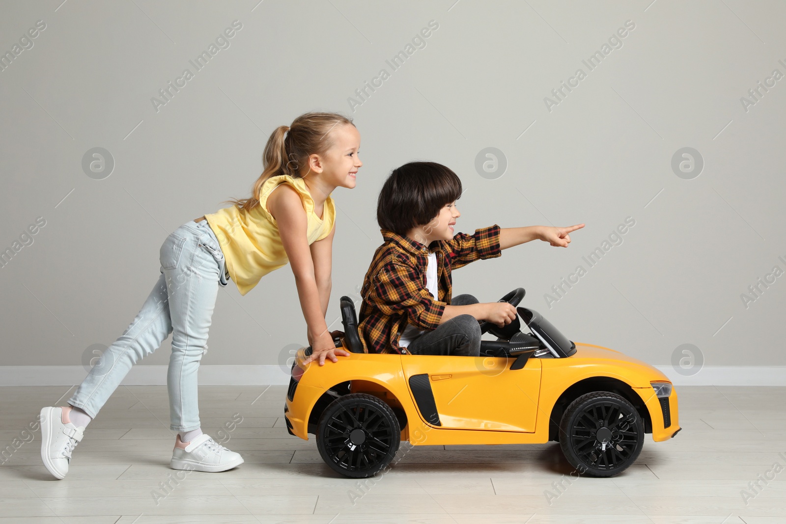 Photo of Cute girl pushing children's electric toy car with little boy near grey wall indoors