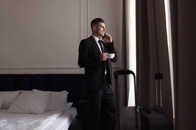 Photo of Handsome businessman drinking coffee while talking on phone near window in hotel room