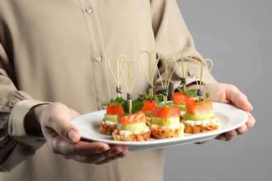 Photo of Woman holding tasty canapes with salmon, cucumber, bread and cream cheese on grey background, closeup