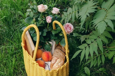 Photo of Yellow wicker bag with roses, peaches, book and baguette on green grass outdoors