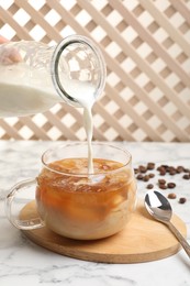 Photo of Woman pouring milk into glass with refreshing iced coffee at white marble table, closeup