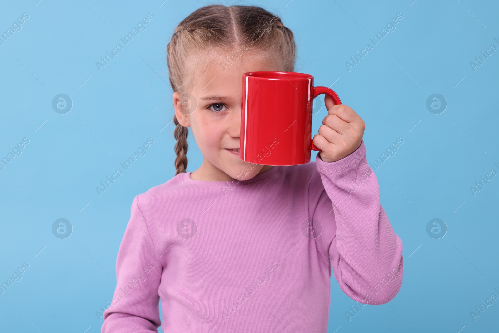 Photo of Cute girl covering eye with red ceramic mug on light blue background