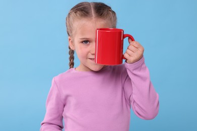 Photo of Cute girl covering eye with red ceramic mug on light blue background