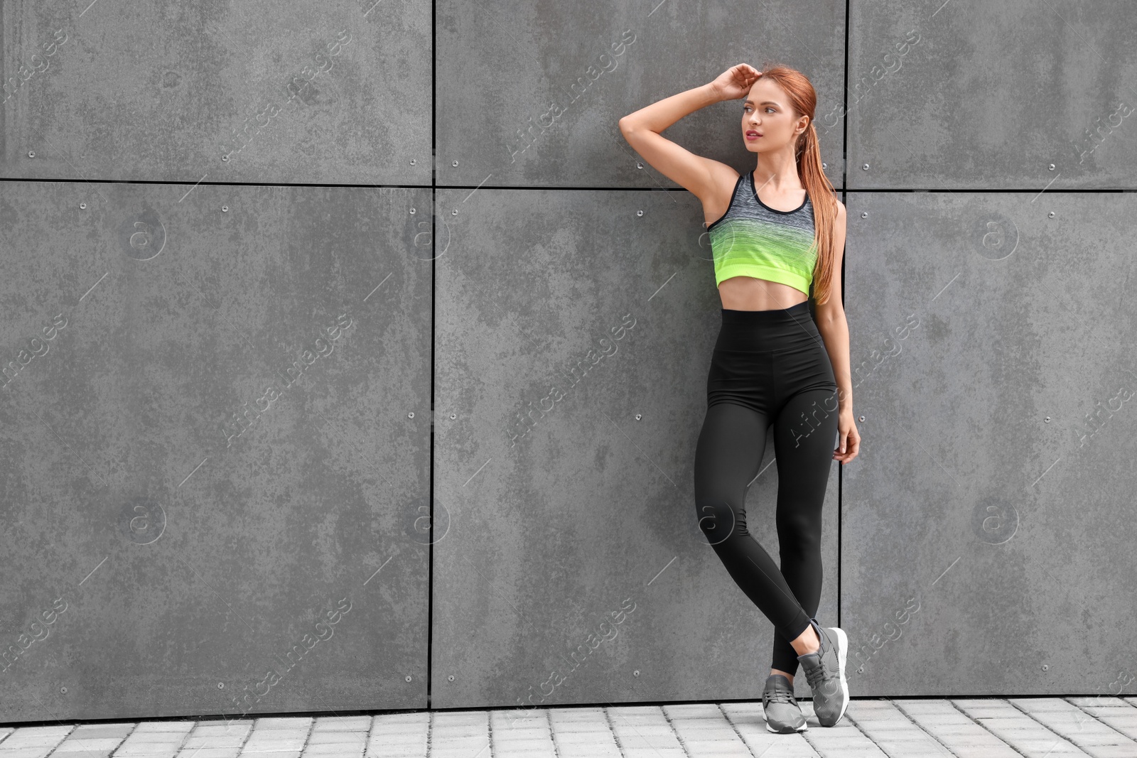 Photo of Beautiful woman in stylish gym clothes posing near grey wall on street, space for text