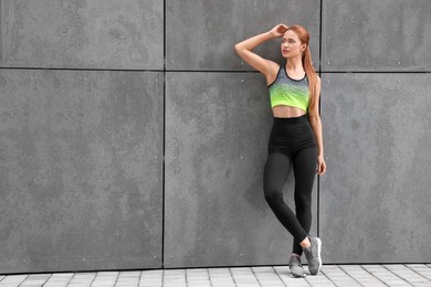 Photo of Beautiful woman in stylish gym clothes posing near grey wall on street, space for text
