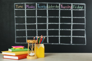 Photo of Hourglass and stationery on white wooden table near blackboard with drawn school timetable
