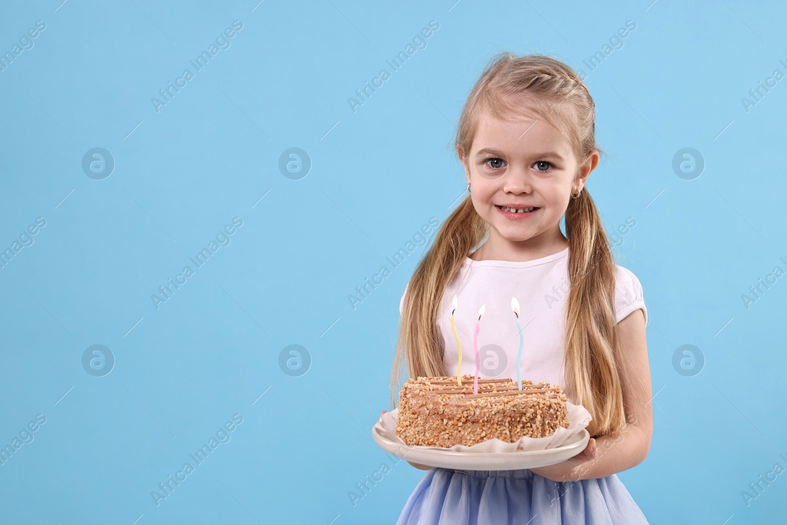 Photo of Birthday celebration. Cute little girl holding tasty cake with burning candles on light blue background, space for text