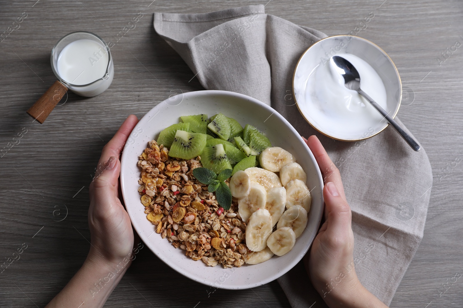 Photo of Woman holding bowl of tasty granola with banana and kiwi at grey wooden table, top view