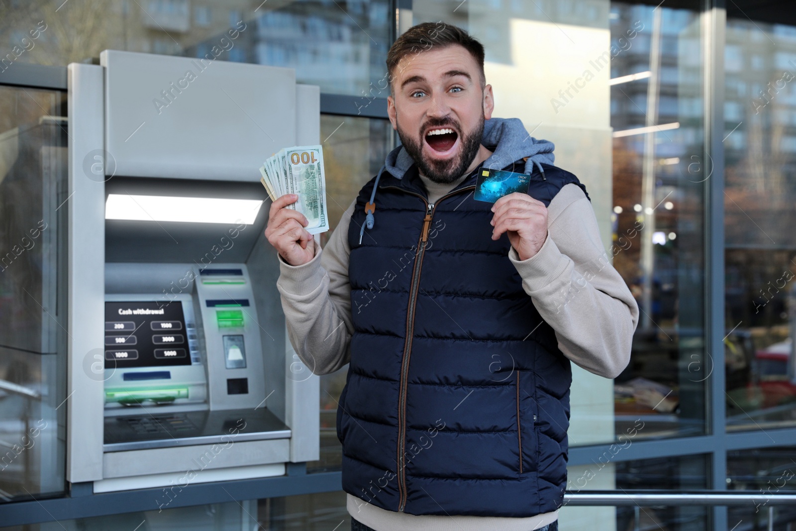 Image of Excited young man with credit card and money near cash machine outdoors
