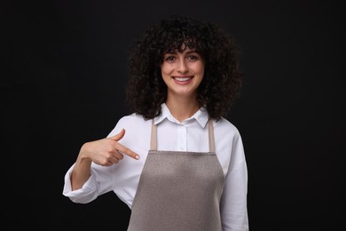 Photo of Happy woman pointing at kitchen apron on black background. Mockup for design
