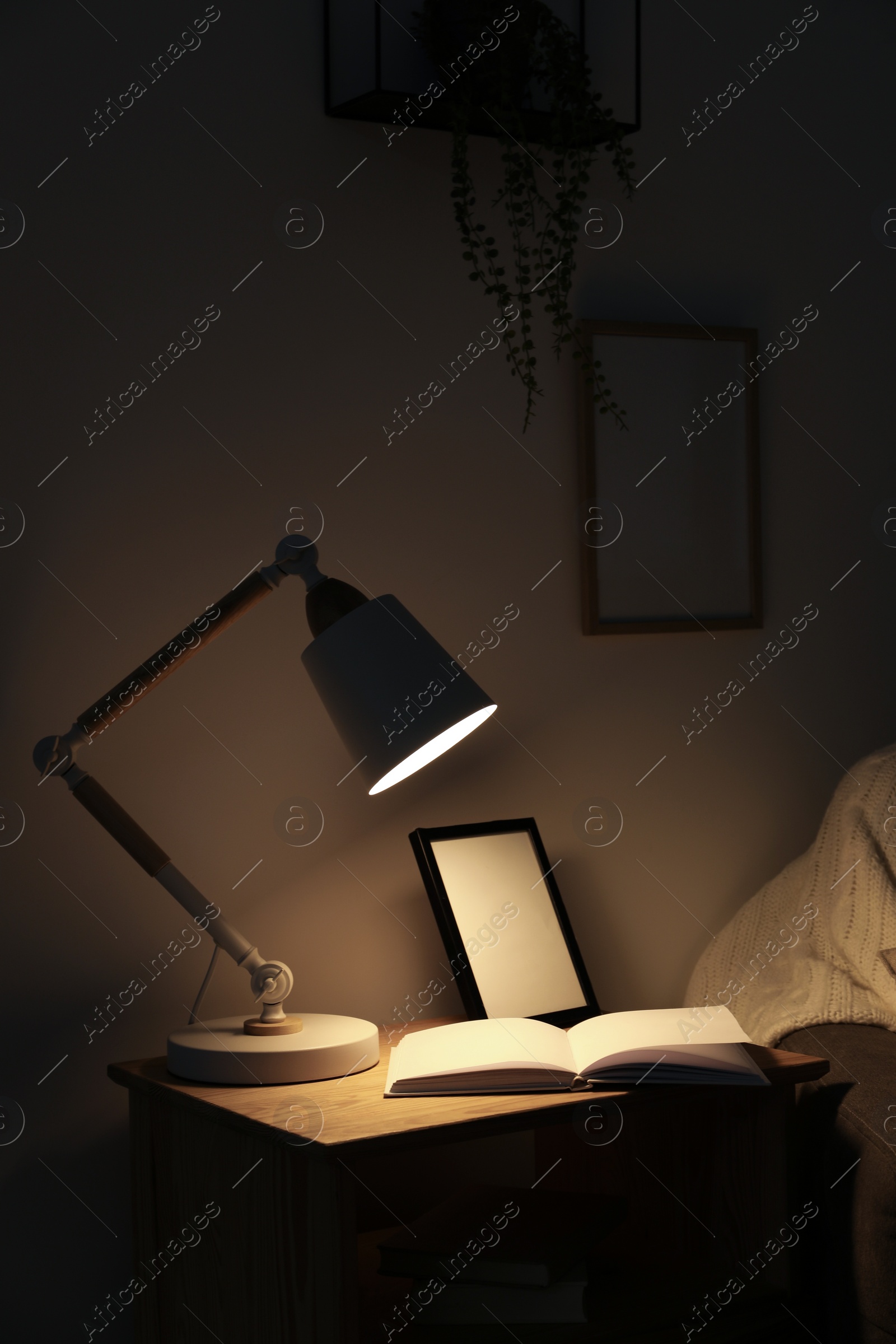 Photo of Stylish modern desk lamp, open book and frame on wooden cabinet in dark room
