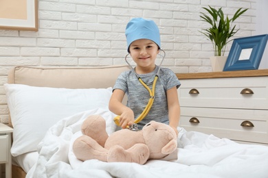 Photo of Cute child playing doctor with stuffed toy on bed in hospital ward