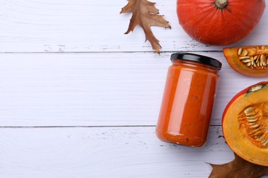 Photo of Jar of delicious pumpkin jam, fresh pumpkins and dry leaves on white wooden table, flat lay. Space for text