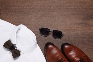 Photo of White shirt with stylish brown bow tie, sunglasses and shoes on wooden background, flat lay. Space for text