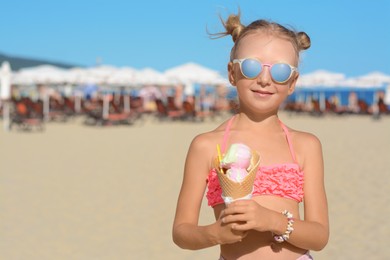 Photo of Adorable little girl in swimsuit with delicious ice cream at beach on sunny summer day, space for text