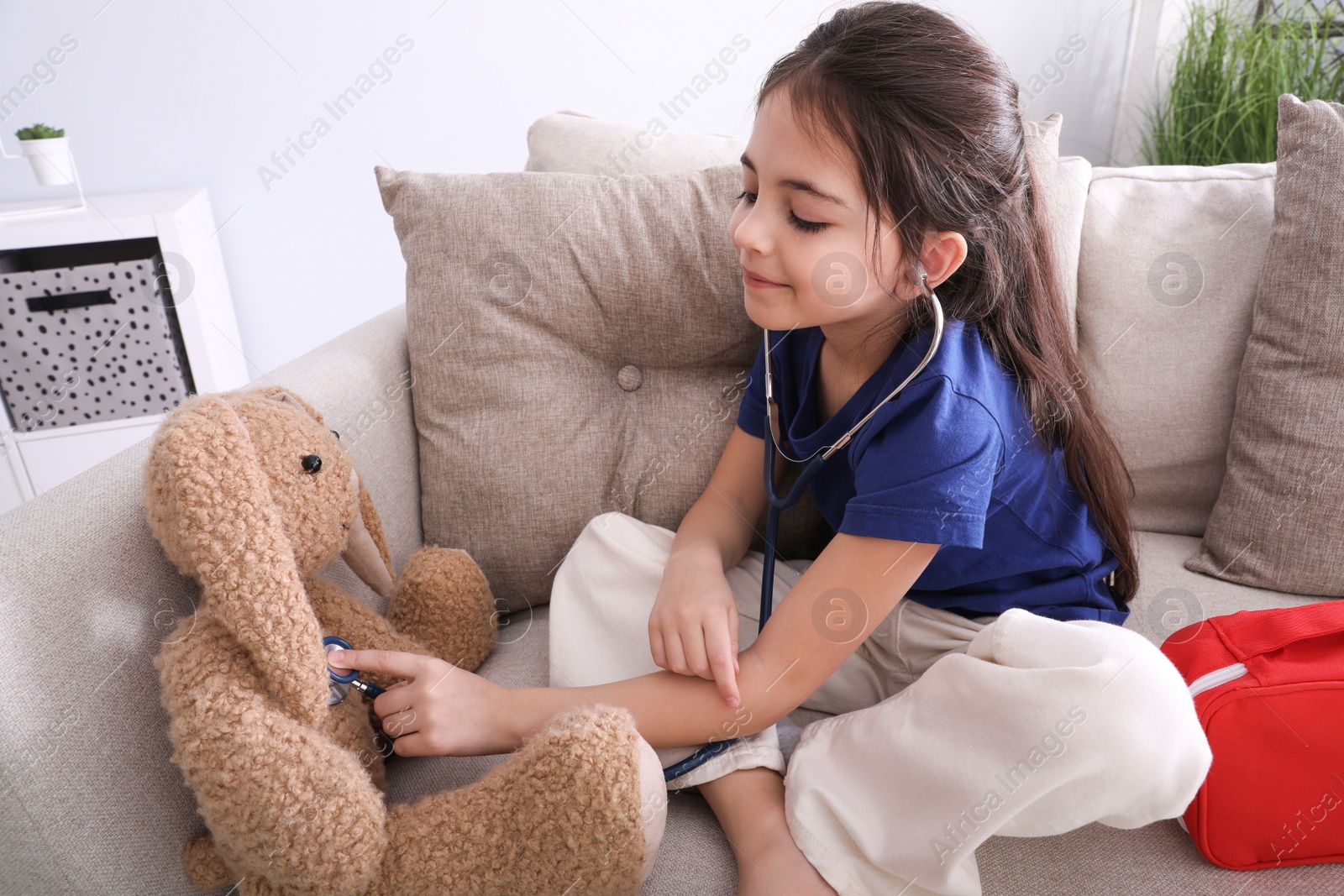 Photo of Little girl playing doctor with toy bunny on sofa indoors. Pediatrician practice