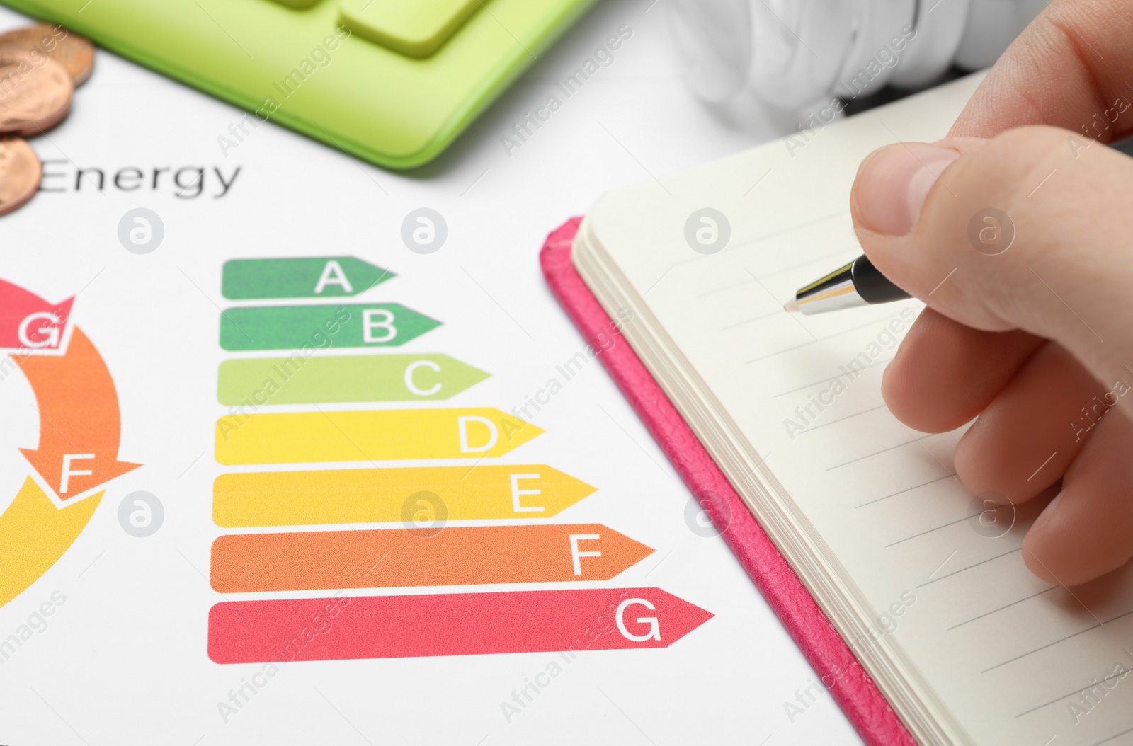 Photo of Woman with pen, notebook, energy efficiency rating chart and coins at table, closeup